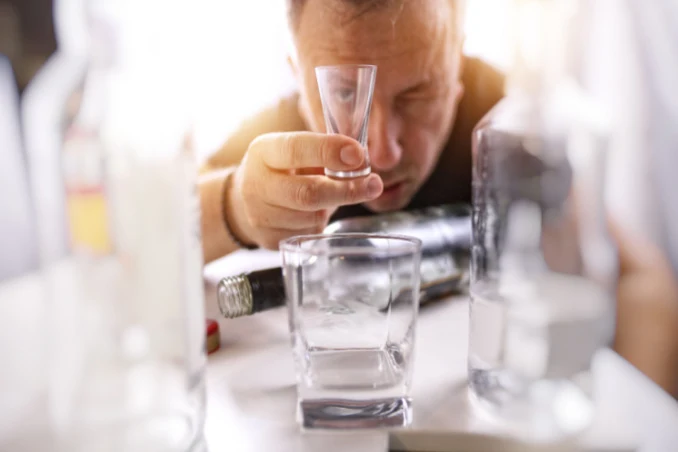 A man surrounded by empty bottles stares through shot glass with the clear need for alcohol detox