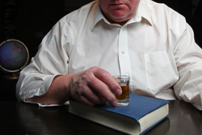 Choosing an AA sponsor can mean the difference between sobriety and drinking, as shown by concept pic of man with a shot glass atop the Big Book