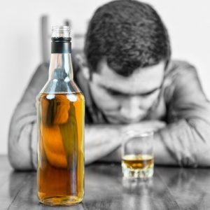 Alcohol Detox - Man with his head down on top of his folded arms on a table with a shot glass and a bottle of alcohol in from of him.