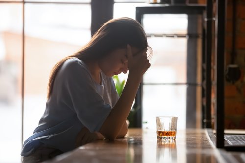 Alcohol and Anxiety - Woman sitting at a bar by herself with her elbow on the bar and her hand on her forehead looking down with a rocks glass of alcohol in front of her.