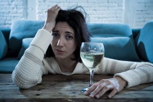 Alcoholism and Depression - Woman sitting in front of her couch with her arms on her coffee table with one hand up to her forehead and the other holding a glass of white wine. She is looking directly at the camera with a depressed look in her eyes.