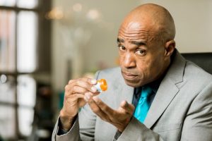 Antidepressant Addiction - Black man in a suit and tie is looking at the camera holding a prescription pill bottle shaking it out into his left hand.
