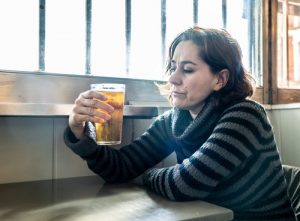 Beer Addiction Rehab, a Woman sitting at a table next to a window in a restaurant drinking a draft beer from a pint glass.
