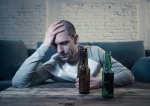 Beer Addiction Symptoms, shown by a man sitting on the floor in front of his couch drinking a beer with 2 empty beer bottles sitting on the coffee table in front of him.