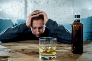 Causes of Alcohol Addiction - man sitting on a couch leaning on a coffee table with his elbows on the table and his hands on top of his heading looking down slightly out of focus. In from of the man in clear focus is a bottle of alcohol and a half empty glass of liquor with some melted ice.