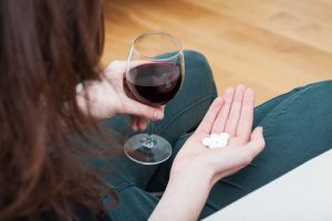 Dual Diagnosis - Photo taken over the right hand shoulder of a woman looking down into her lap, holding a glass of red wine in one hand and several white pills in the other hand.
