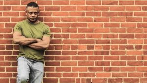 PTSD and Drugs - Man standing, leaning against a brick wall with arms crossed in front of him looking down at the ground.