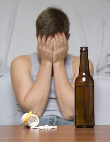 Addiction - Man in a tank top sits in front of a couch with both hands held over his face. A bottle of pills is overturned on a table in front of him with several pills spilling out and a bottle to beer. He is dealing with addiction.