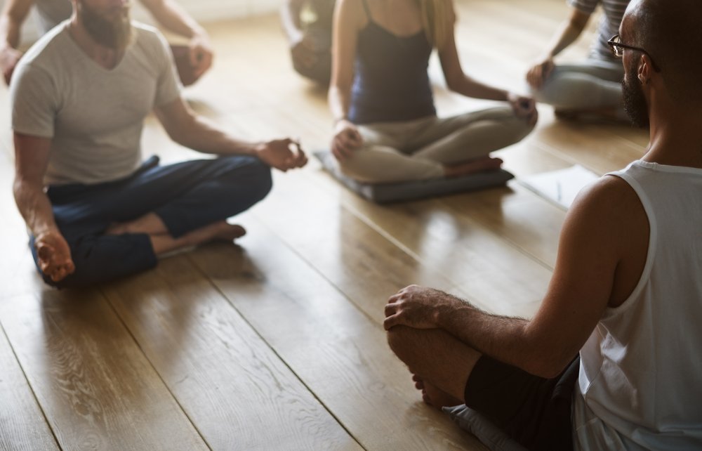 Addiction Recovery - Photo of several people using meditation in addiction recovery. You see serval people from the neck down siting on the floor, legs crossed with their hands in a meditation pose as the face the instructor.