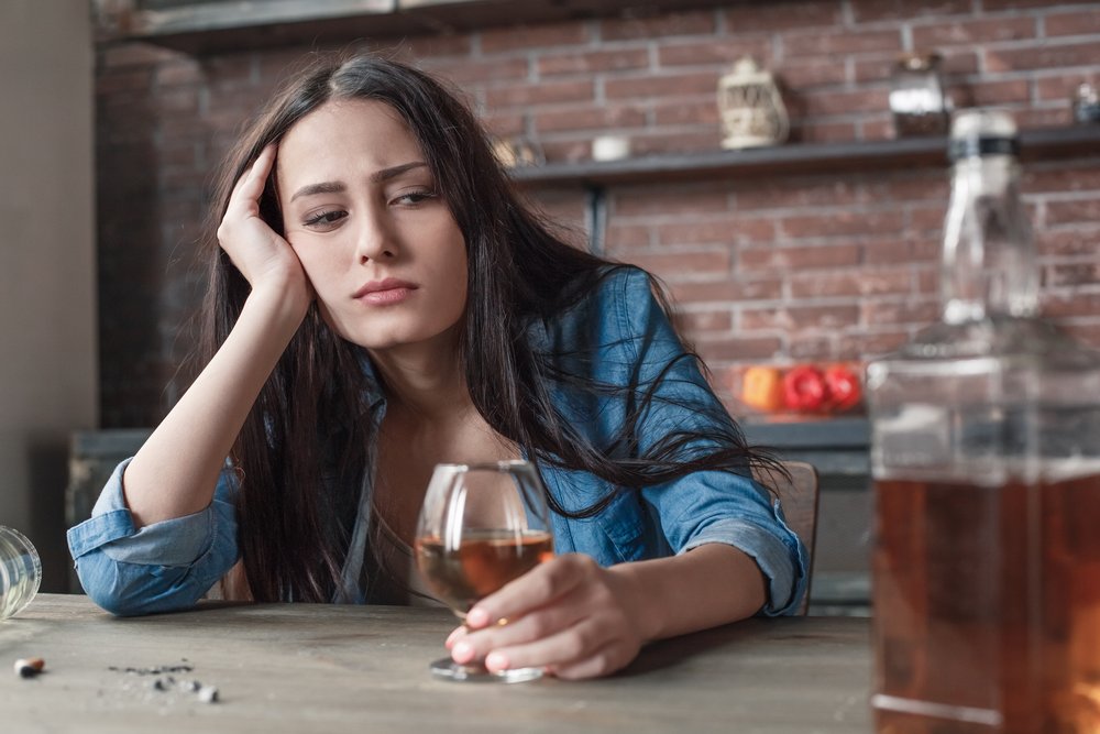 Alcoholism - A young female alcoholic sits at a table drinking brandy alone.