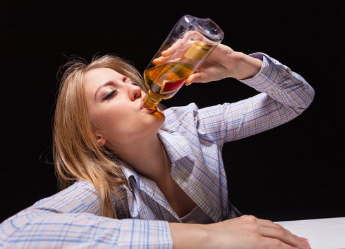 Alcoholism - Photo of a woman sitting at a table drinking straight from a bottle of scotch. To show the concept of How to Tell if Someone is an Alcoholic