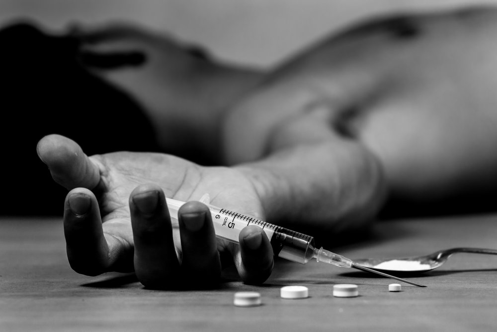 Drug Overdose- black and white photo of a man suffering from a drug overdose, laying down on the ground with his head turned away from the camera. His arm is stretched out towards the camera with a drug syringe in his open hand. 4 pills and a spoon with white powder are next to his hand. To show concept of what to do when someone overdoses