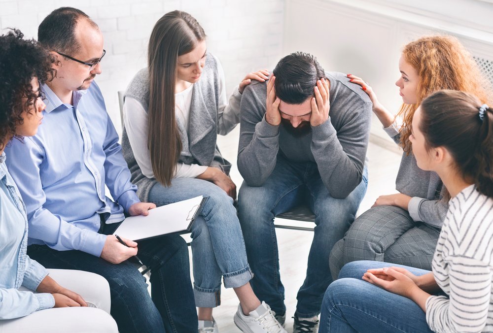 Group Therapy - A man sits in a circle with a group of 4 women and another man. He has his head in his hands as the women on either side console him by placing their hands on his shoulders during group therapy for addiction treatment.