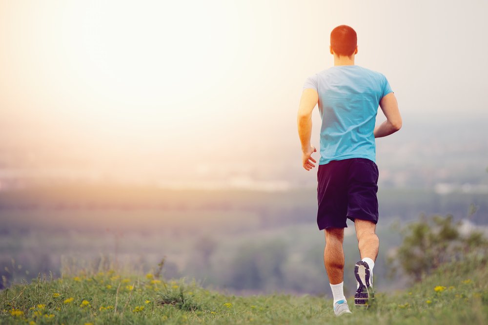 Healthy in Sobriety - Photo from behind a man running in a flowery meadow with the sun low on the horizon shining at him over looking a green valley. The man is exercising staying healthy in sobriety.