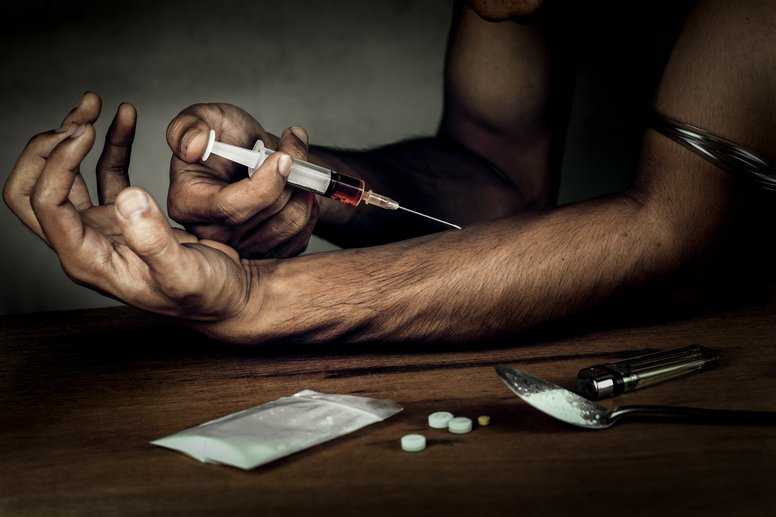 Heroin Addiction - Dark image of a man holding a heroin needle in his right hand injection it into a vein on his left arm.