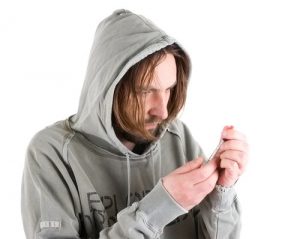 Heroin Addiction Signs - Rough looking Man with long hair in a dirty, ratty hoodie holding a needle wit heroin in it.