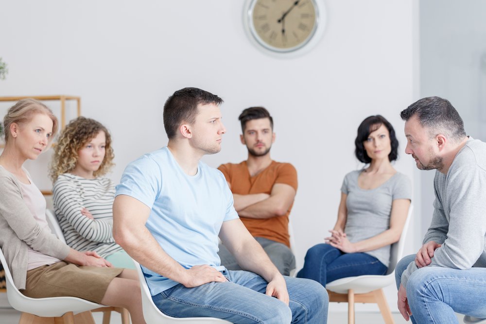 To show an IOP program, a group of 6 people meet for their IOP therapy for addiction treatment. In the foreground 2 men look at each other with serious faces. In the background 3 women and a man look on as the two men talk to each other.