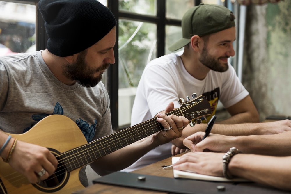 To show Music Therapy, a photo of 2 men sitting on one side of a table. The man closest to the camera is playing a guitar the man next to him sits smiling looking across the table. From the other side of the table you see a pair of hands writing with a pen on a sheet of paper during the music therapy during addiction treatment.