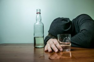 Myths About Alcoholism - mans sits at a table with his head down covered by a hoodie. he as a drink in is right hand and a half full bottle of alcohol sits next time him. Looks like he is trying to deal with alcoholism.