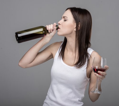 Quit Drinking Alcohol - Middle aged woman in a white tank top with a glass of wine in her left hand and drinking from a bottle of wine in her right hand. She needs help to quit drinking alcohol.