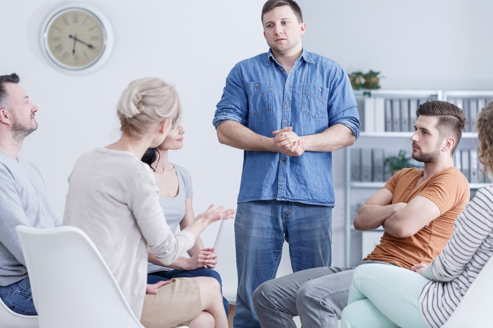 Rehab - Man stands up during group therapy in addiction rehab to talk about his issues. He is wearing a jean shirt and blue jeans standing in the middle of 2 men and 3 women who are seated listening to him and the therapist.