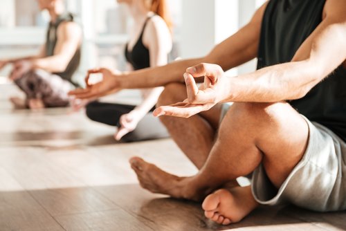 Staying Sober - Photo of a man sitting with his legs crossed on the floor with both forearms resting on his knees with both pointer fingers touching his thumbs in a yoga pose. In the back ground there is a woman who is slightly blurry sitting in the same pose and then another man just past her in the same pose also.