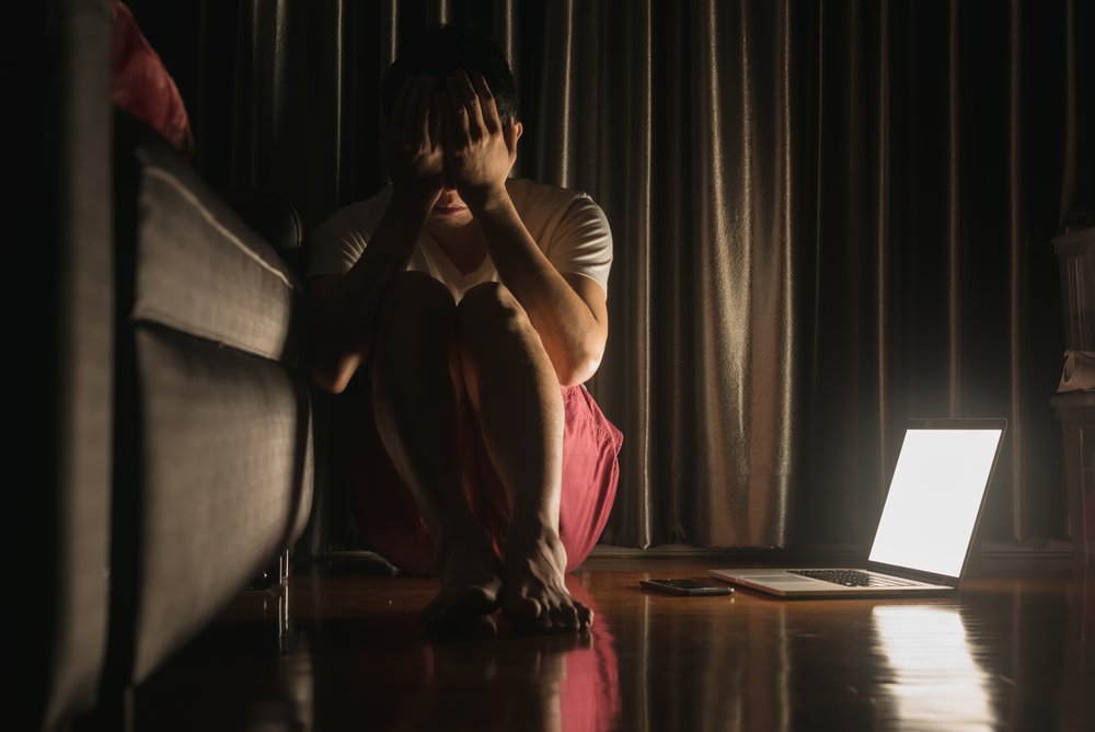 A woman sits on the floor next to her couch in a dark room. The light from her laptop lights up the space, to illustrate the potential power of art therapy for recovery within reach