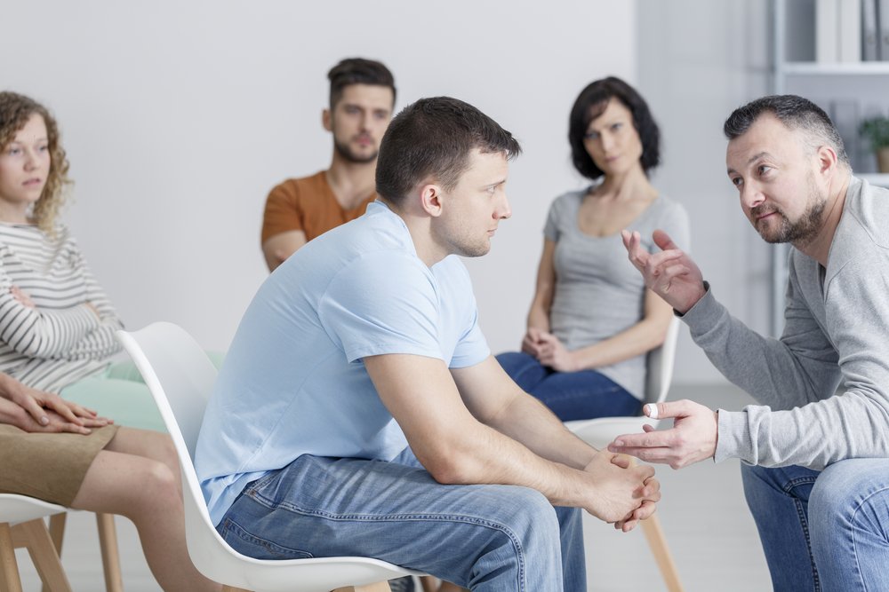 Balance in Sobriety - A group of people sit in a meeting for recovering addicts as 2 men look at teach other and discuss how to maintain balance in sobriety.