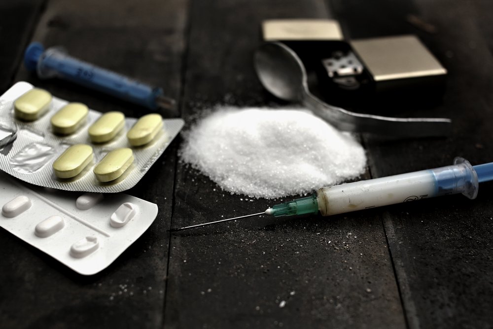 Heroin - A heroin needle with a spoon and lighter sit next to some powder and a couple of packs of pills.