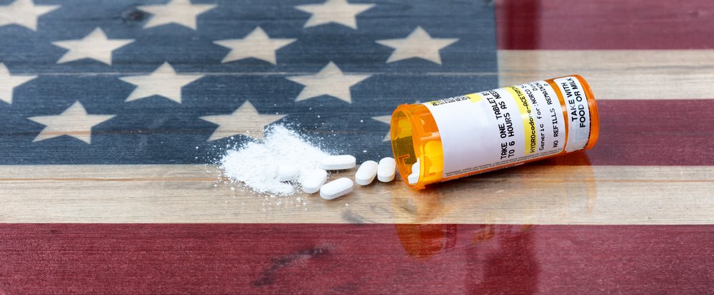 Opioid Epidemic - Photo of a pill bottle spilled out with some whole pills and some crushed into a powder laying on top of a wooden American flag.