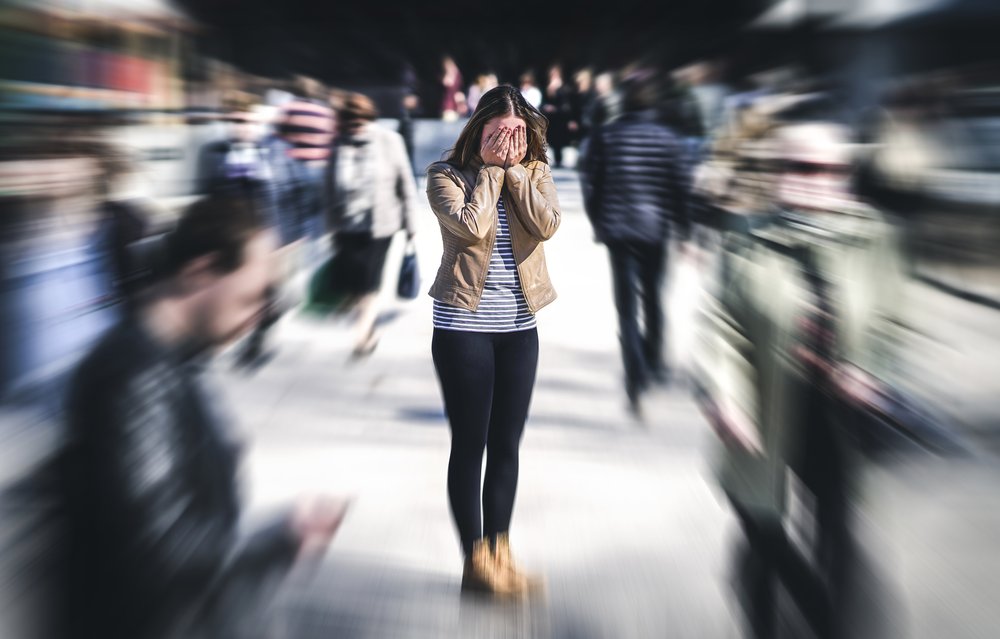 Panic Attack - Photo of a woman standing in the middle of a very busy area with several people walking in different directions around her. She in in focus in the middle of the photo as everyone else is blurry from movement simulating a panic attack.