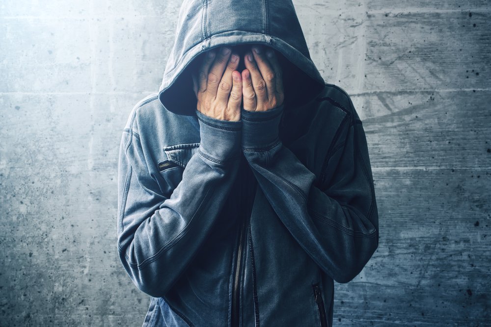 Sobriety - A young man in a grey jacket with the hood over his head who recently completed addiction treatment, stands against a grey wall with his head down and his hands covering his face. He is overwhelmed with just being in society and maintaining his sobriety.
