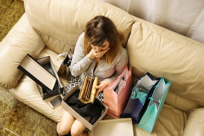 Overhead view: a woman sits with an empty wallet on her couch, surrounded by shopping bags