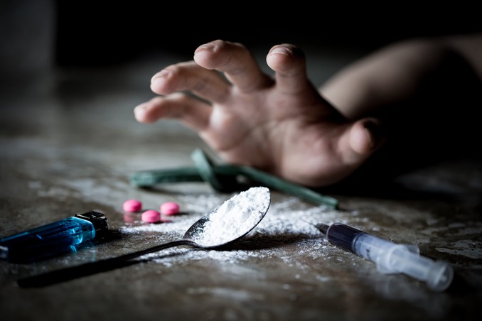 A hand reaches out for a drug spoon, amongst drug paraphernalia and powders