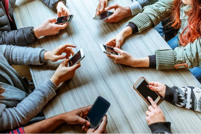 Close view of six people's hands at a table, all holding cell phones and using social media 